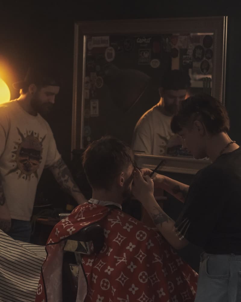 Barberka goli klienta w fotelu, za nimi drugi barber przy lustrze w ciepłym świetle lampy